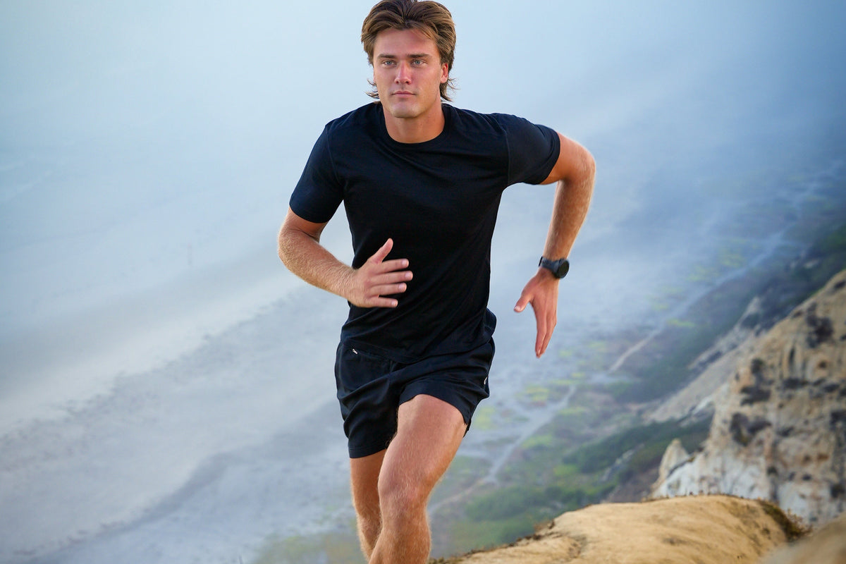 A male runner in a black athletic t-shirt and black shorts is captured mid-stride on a dirt trail with a misty mountain landscape in the background.